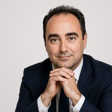 Portrait de Romain Duinat, souriant, en costume sombre, les mains jointes